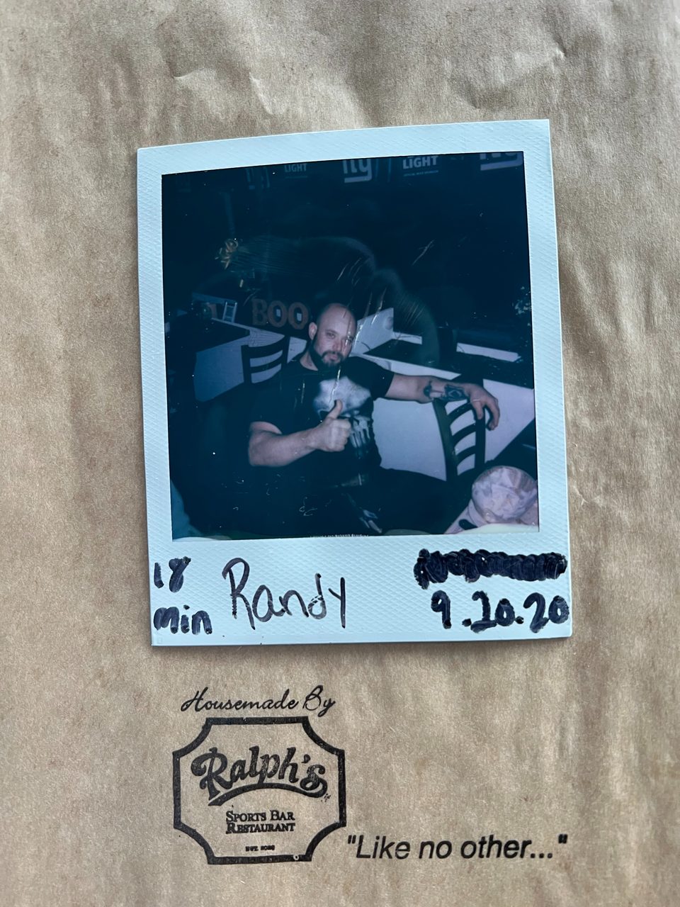 A Polaroid photo of a man giving a thumbs-up, with "Randy 9.10.20" written below, on a Ralph's Sports Bar bag.