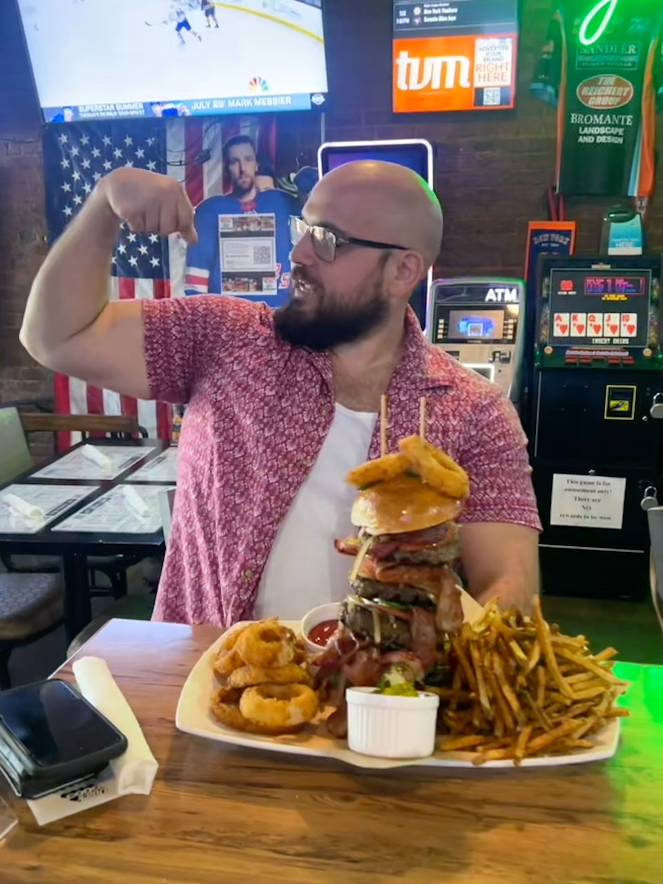 Man flexing his arm next to a giant burger, onion rings, and fries at a bar.