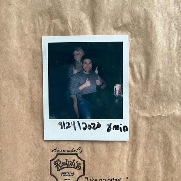 A polaroid of two smiling people giving thumbs up, on a brown paper bag from Ralph's Sports Bar & Restaurant.