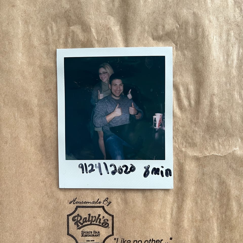 A polaroid of two smiling people giving thumbs up, on a brown paper bag from Ralph's Sports Bar & Restaurant.