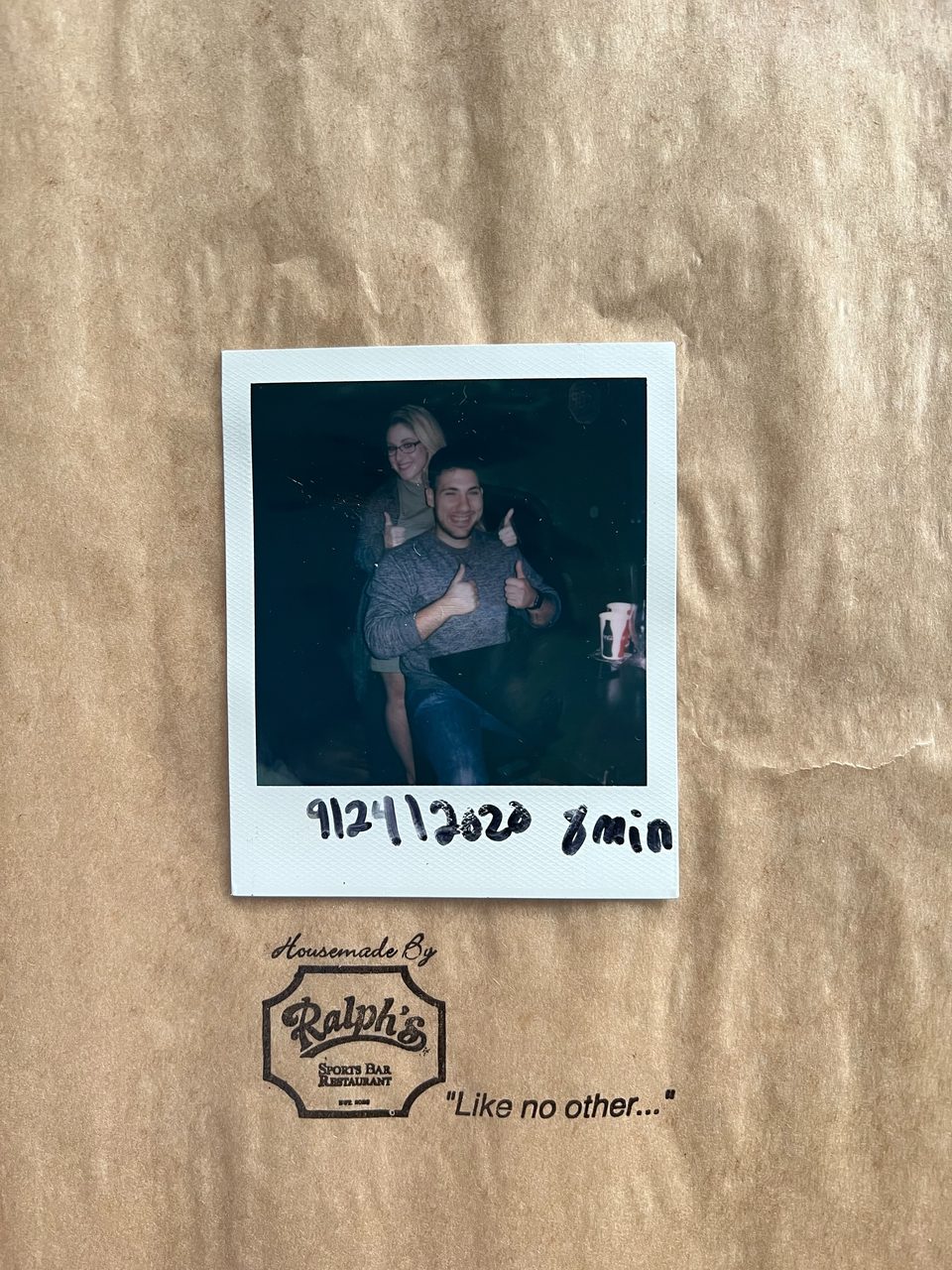 A polaroid of two smiling people giving thumbs up, on a brown paper bag from Ralph's Sports Bar & Restaurant.