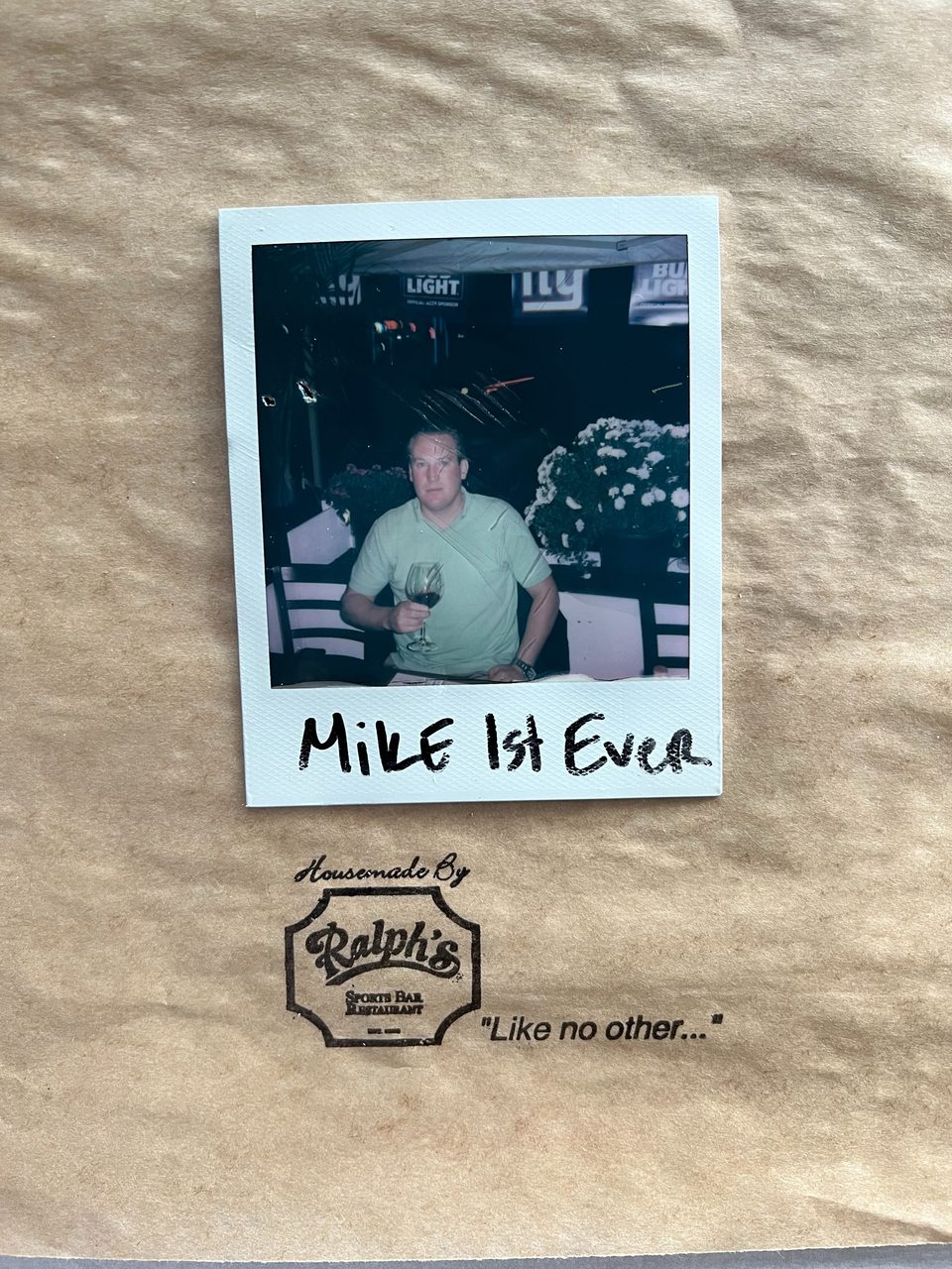 A polaroid of a man in a green shirt holding a glass of red wine, with "Mike 1st Ever" written below.