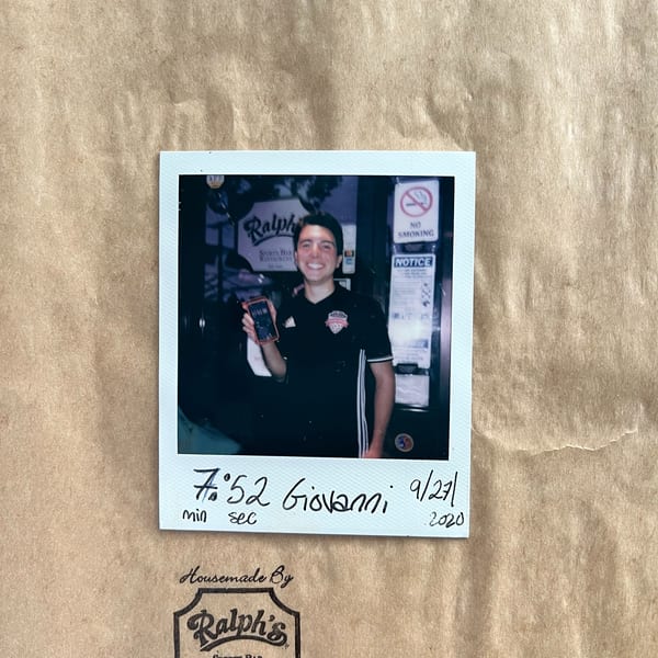A smiling man in a Ralph's Sports Bar shirt holds a phone in a polaroid photo on a brown paper bag.