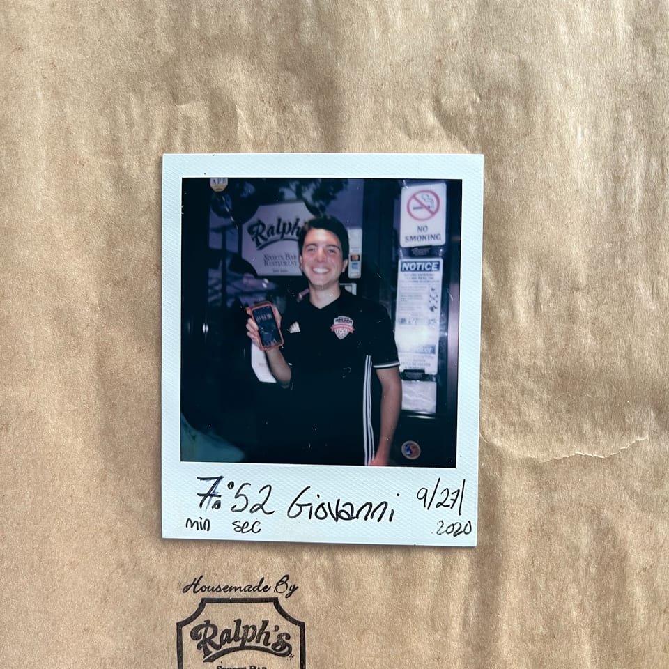 A smiling man in a Ralph's Sports Bar shirt holds a phone in a polaroid photo on a brown paper bag.
