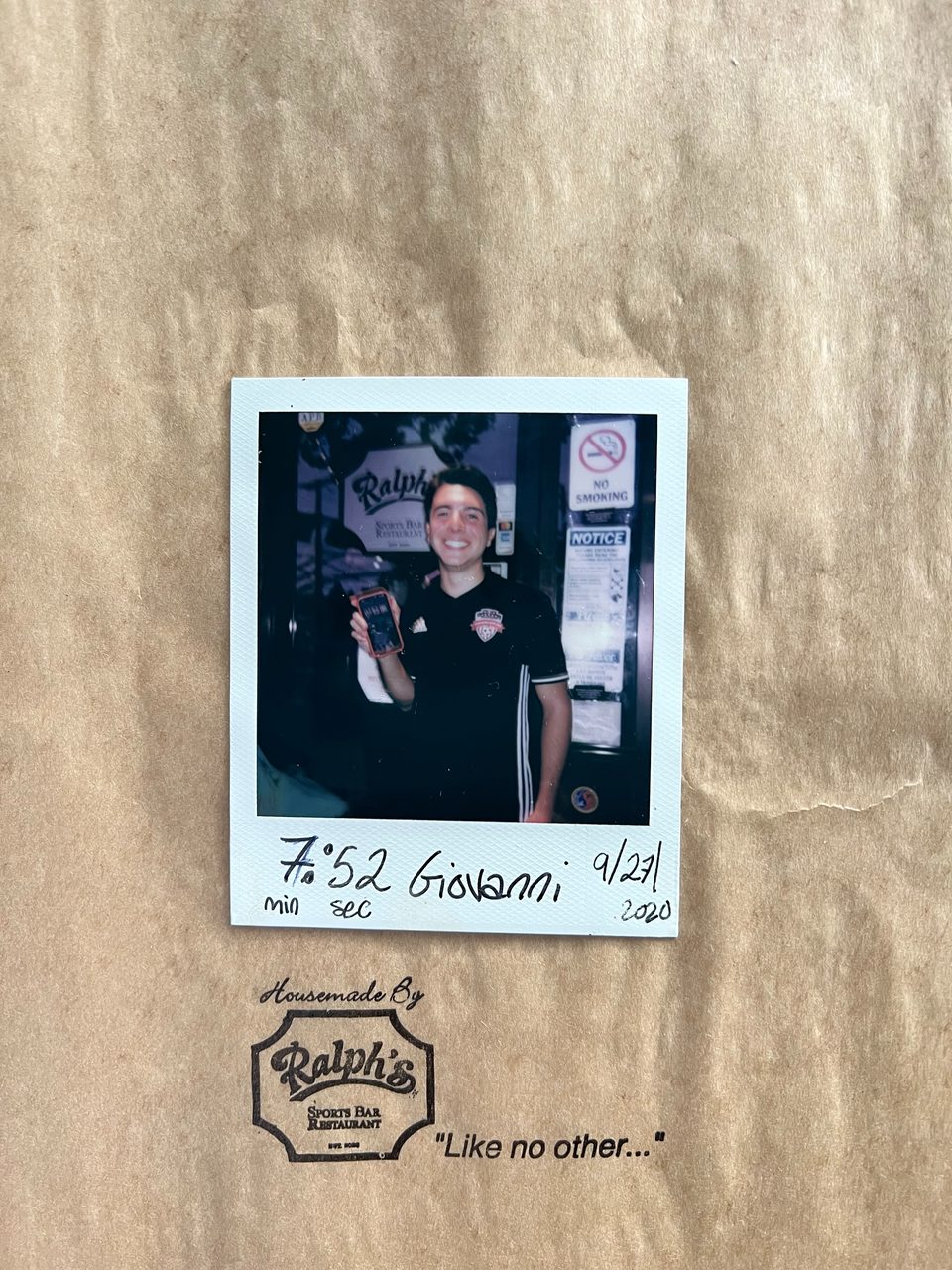A smiling man in a Ralph's Sports Bar shirt holds a phone in a polaroid photo on a brown paper bag.
