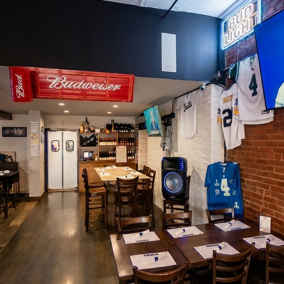 A sports bar interior with a long wooden bar, dining tables, and sports jerseys on brick walls.