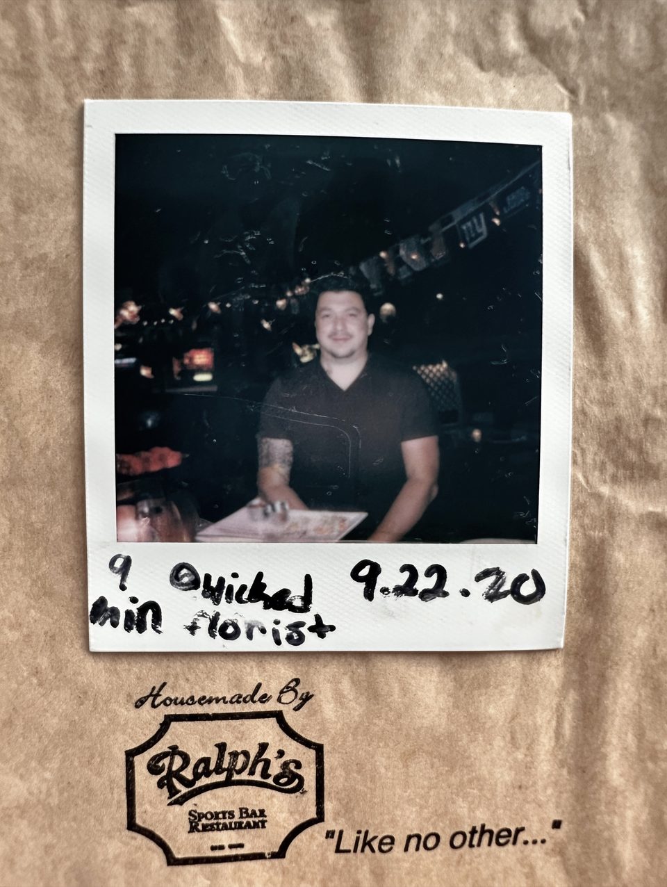 Polaroid of a man with tattoos on his arms, sitting at a table in a dimly lit restaurant.